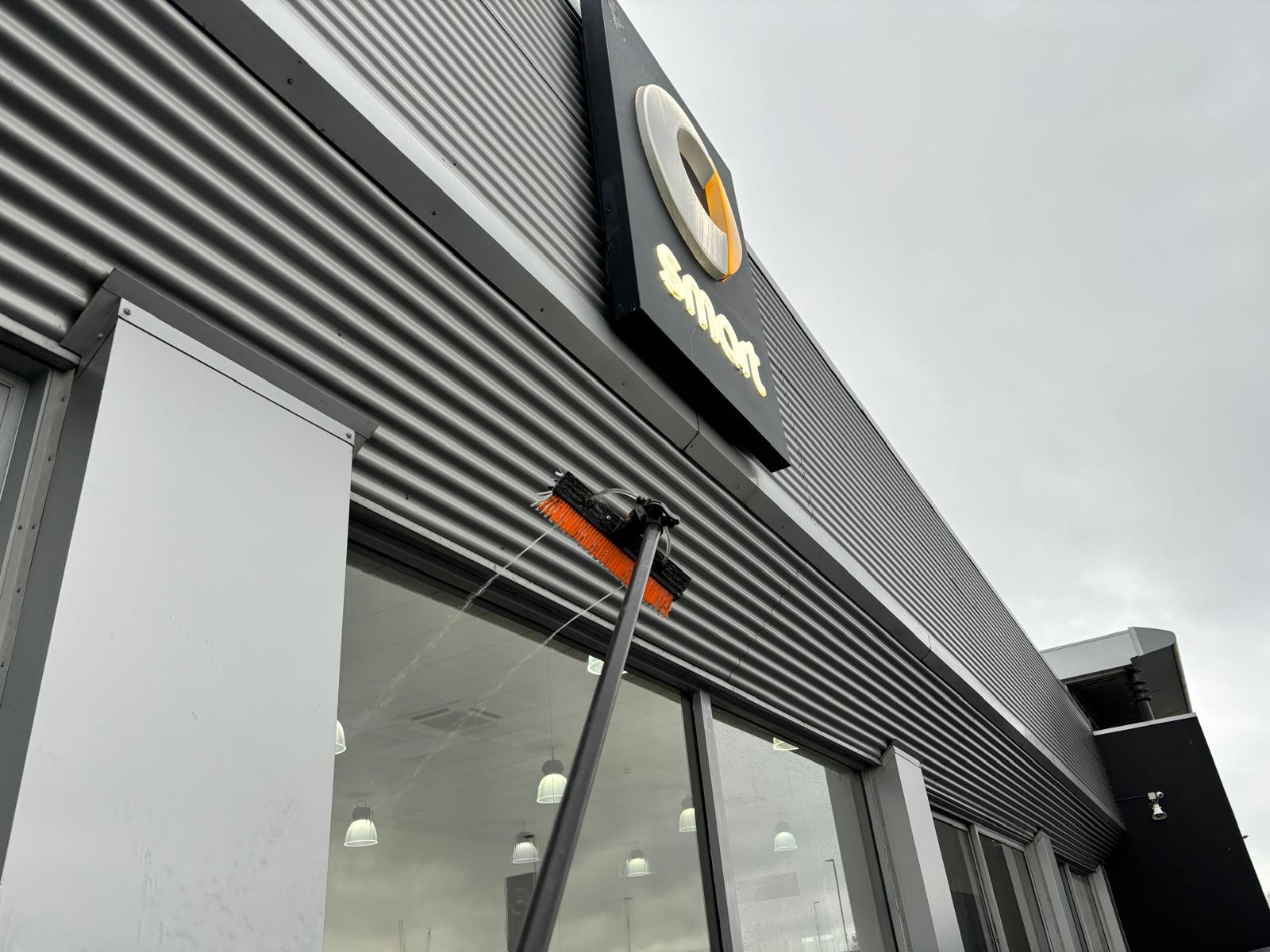 Water-fed pole brush cleaning upper showroom windows beneath the smart dealership sign