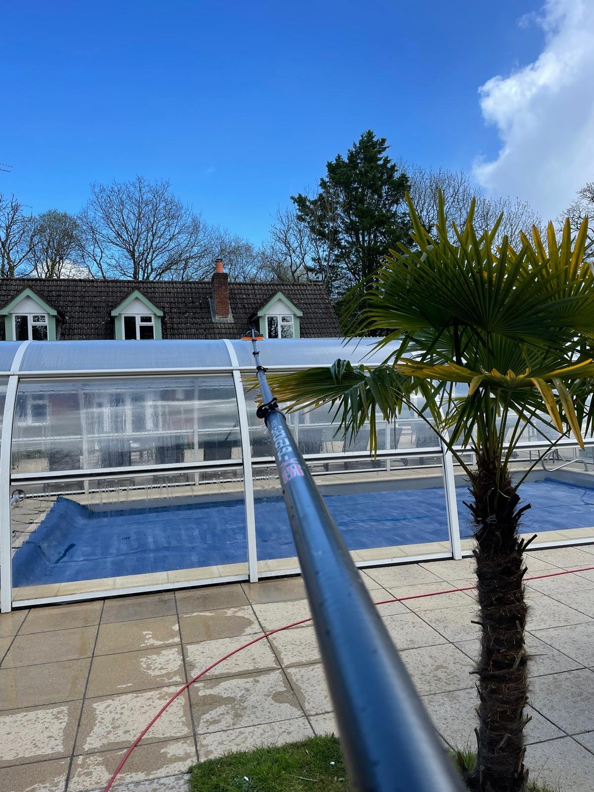 Wide view of a swimming pool enclosure being cleaned with a water-fed pole, house and palm tree in view