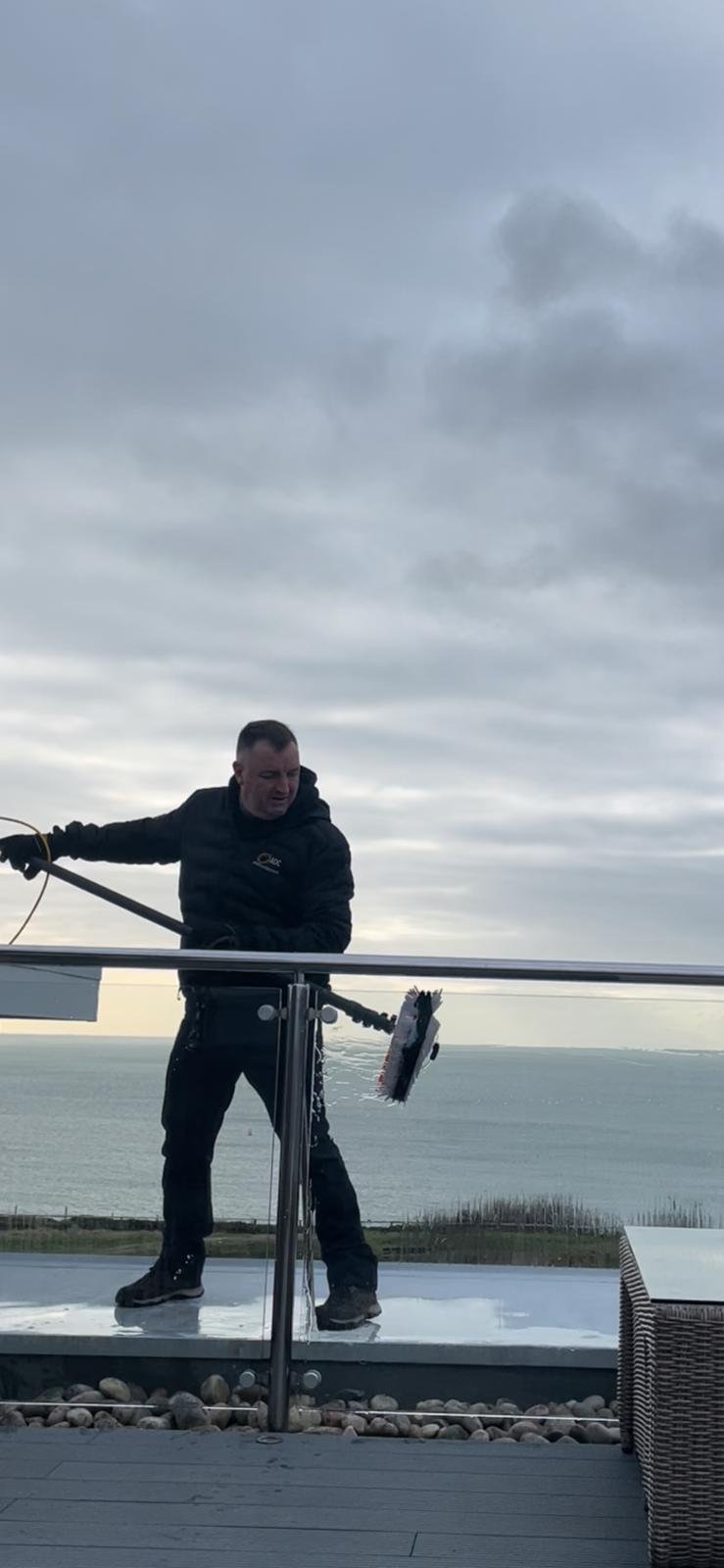 Andy cleaning a rooftop glass balustrade with a water-fed brush, the sea behind him